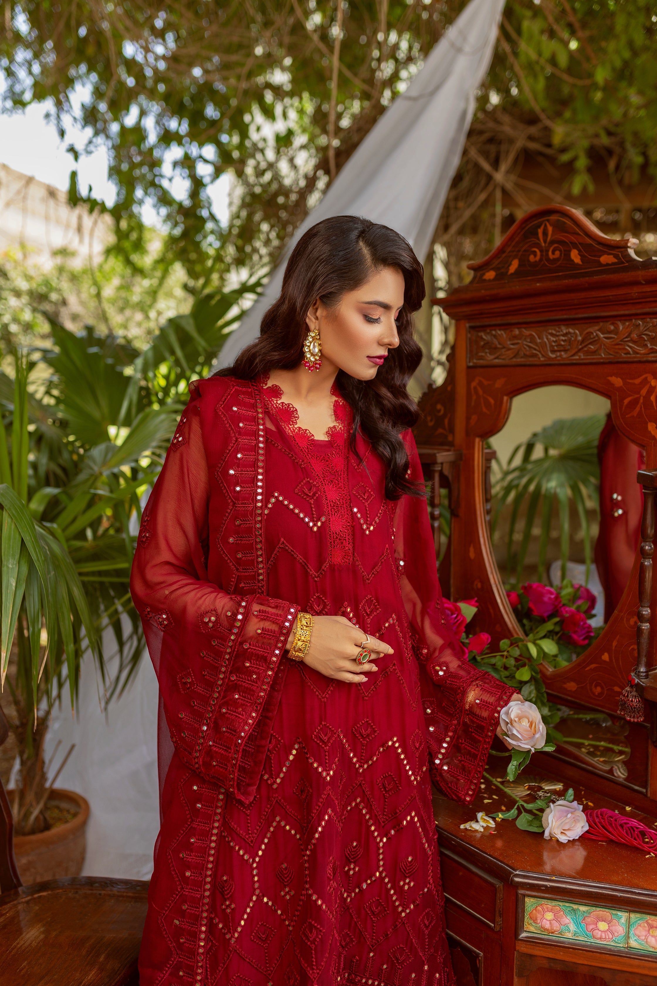 A girl in red embroided chiffon suit standing in front of a mirror with a pink rose in hand.