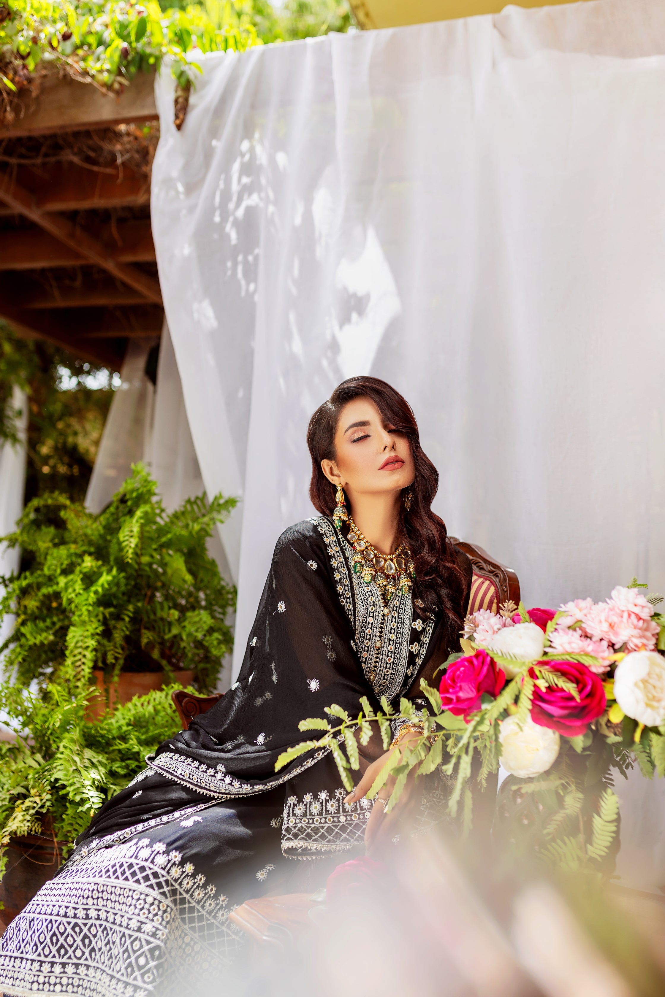 A girl is sitting on a chair in black chiffon dress with a white curtain behind her.