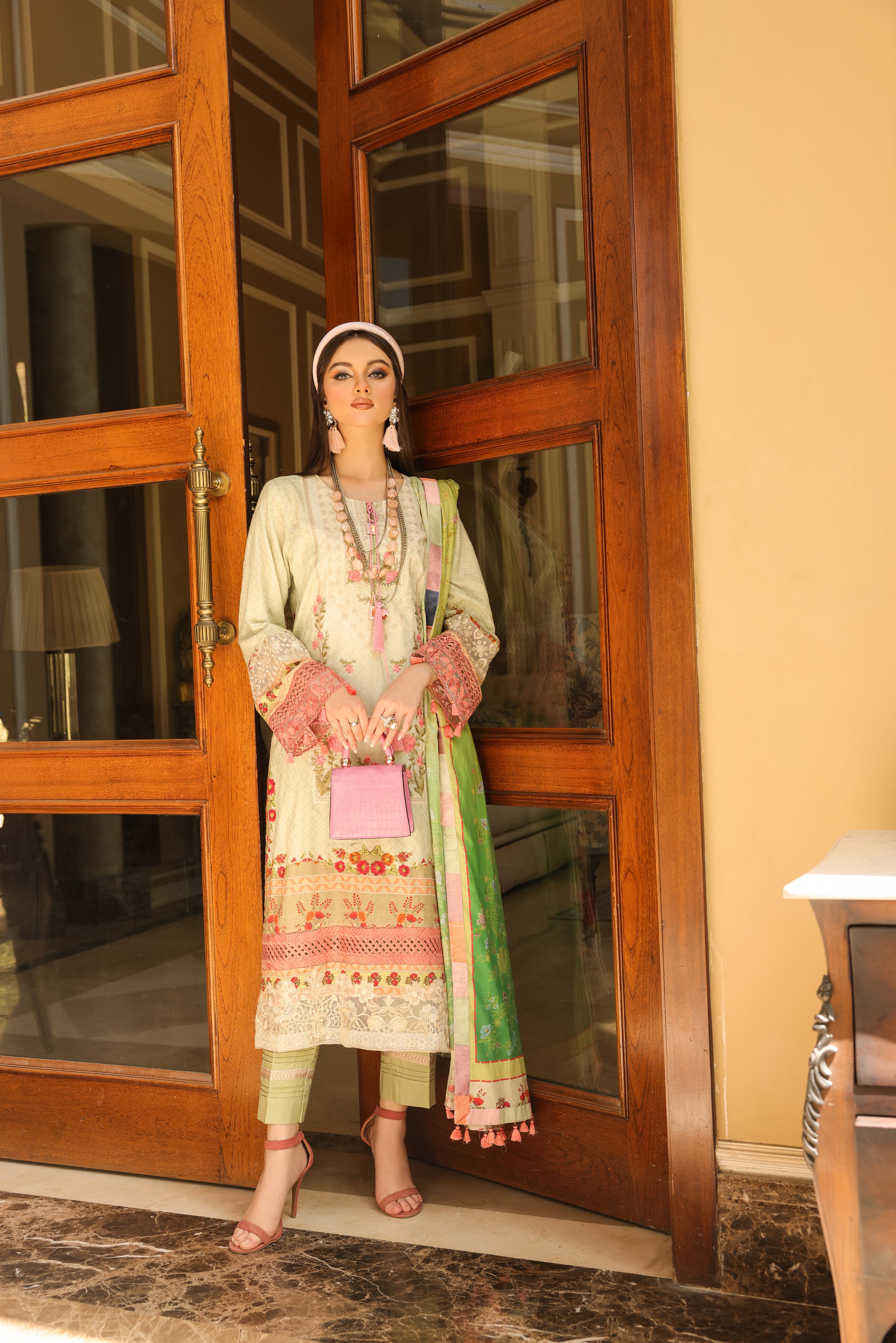 A girl is standing  in a door wearing Embroidered Organza Suit with pink color hand bag.